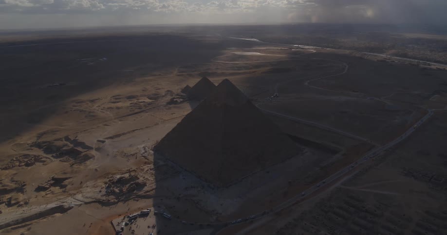 A dramatic high-angle static shot captures the Great Pyramids of Giza casting a long shadow across the vast Egyptian desert