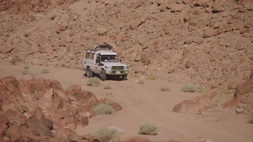 A white off-road safari vehicle slowly drives up a sandy path through a rugged desert canyon