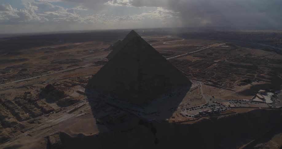 An epic aerial view of the Great Pyramids of Giza standing majestically in the desert under a dramatic, cloudy sky with sun rays shining through
