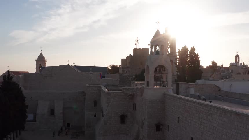 An aerial view slowly pulls back from the Church of the Nativity in Bethlehem, silhouetted against the bright, warm glow of the sun