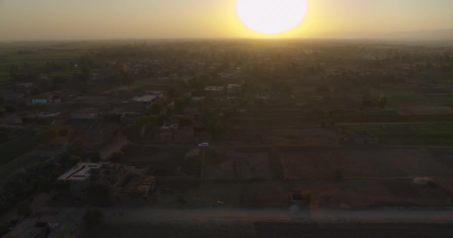 An expansive aerial shot slowly pulls back to reveal a sprawling rural village and farmlands bathed in the golden light of the setting sun