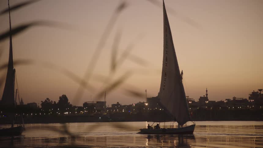 A slow focus pull reveals a traditional felucca sailboat cruising on the Nile river at sunset, with a silhouetted cityscape in the background