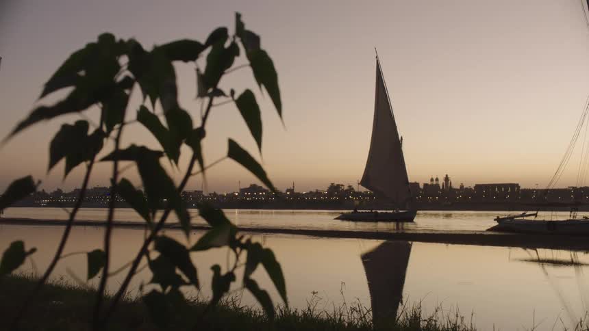 A tranquil view of a traditional felucca sailboat on the calm Nile river at dusk with the city of Luxor in the background