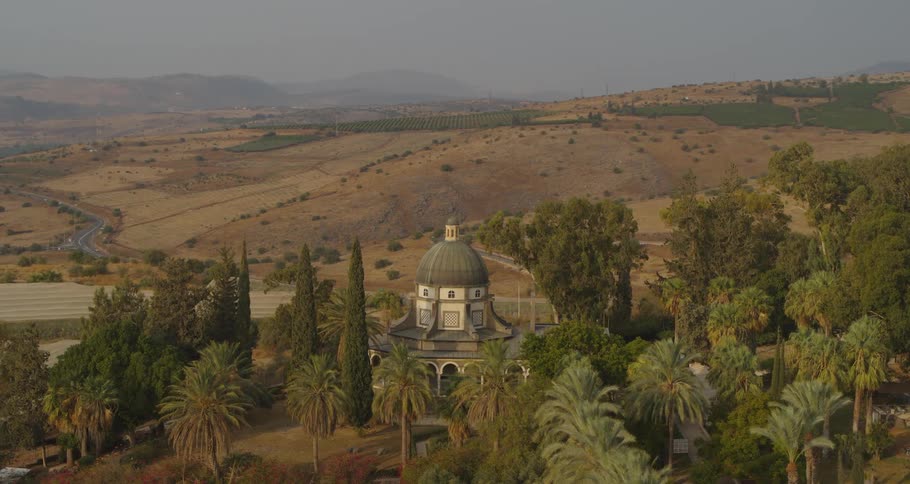 A scenic high-angle view of the Church of the Beatitudes surrounded by lush gardens, with the arid hills of Galilee in the background