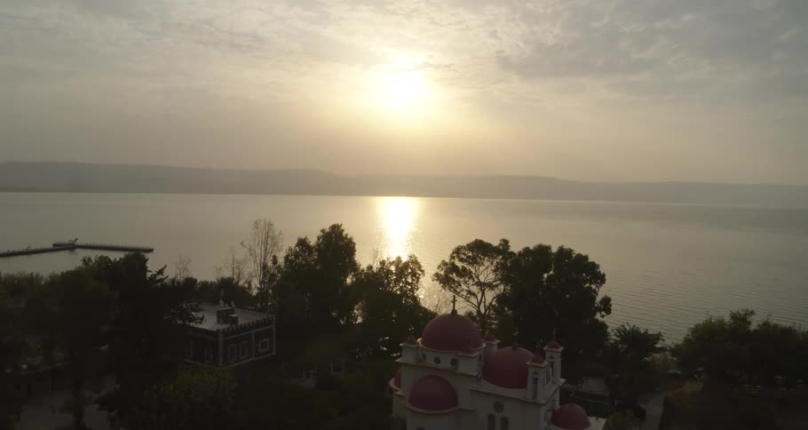 A slow aerial pullback reveals the iconic Greek Orthodox Church with its red domes overlooking the Sea of Galilee at sunrise