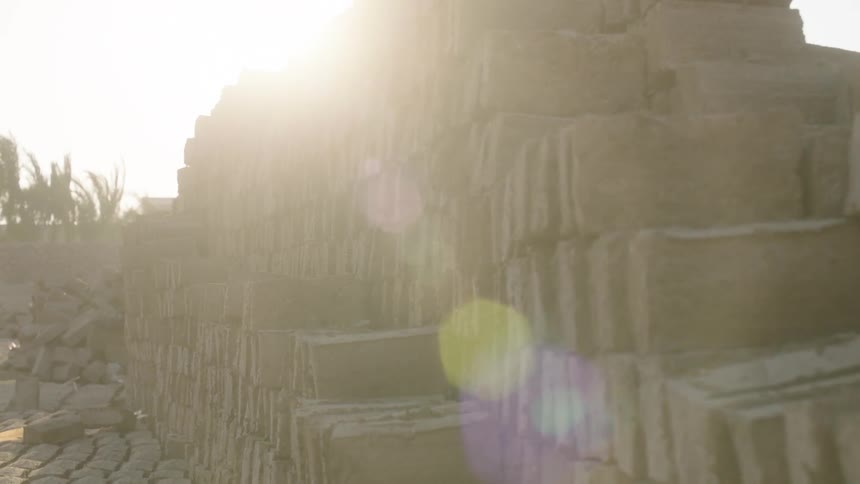 Handmade mud bricks are stacked to dry in the sun, with a beautiful lens flare moving across the frame as the camera pulls back