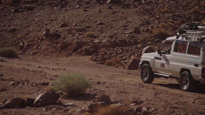 A white off-road safari vehicle is parked on a dusty road in a rugged and rocky desert landscape