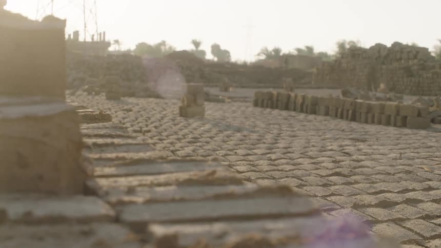 A dolly shot reveals thousands of handmade mud bricks drying in neat rows under the hot sun at a rural brick factory