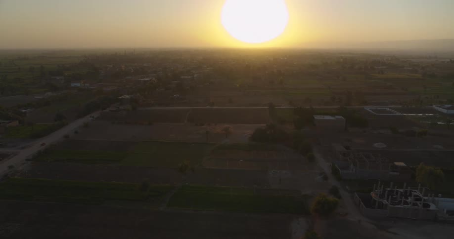 A slow aerial pullback reveals a vast rural landscape of farmlands and a small village under the golden light of a hazy setting sun