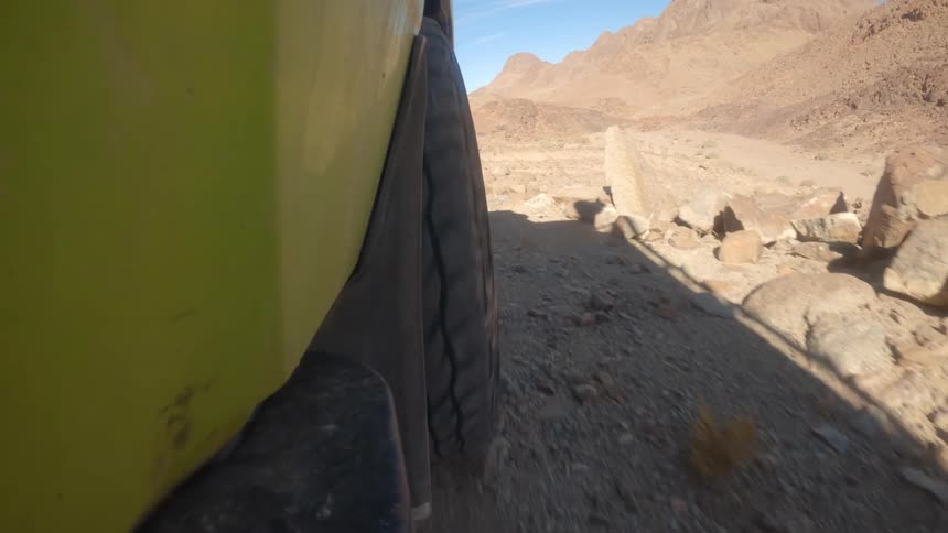 A slow-motion, low-angle shot captures an off-road vehicle's wheel carefully navigating a rugged, rocky trail through a sun-drenched desert