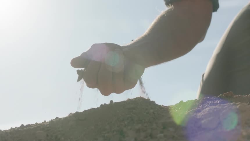 A person's hand lets dry soil sift through their fingers in slow motion, backlit by the bright sun