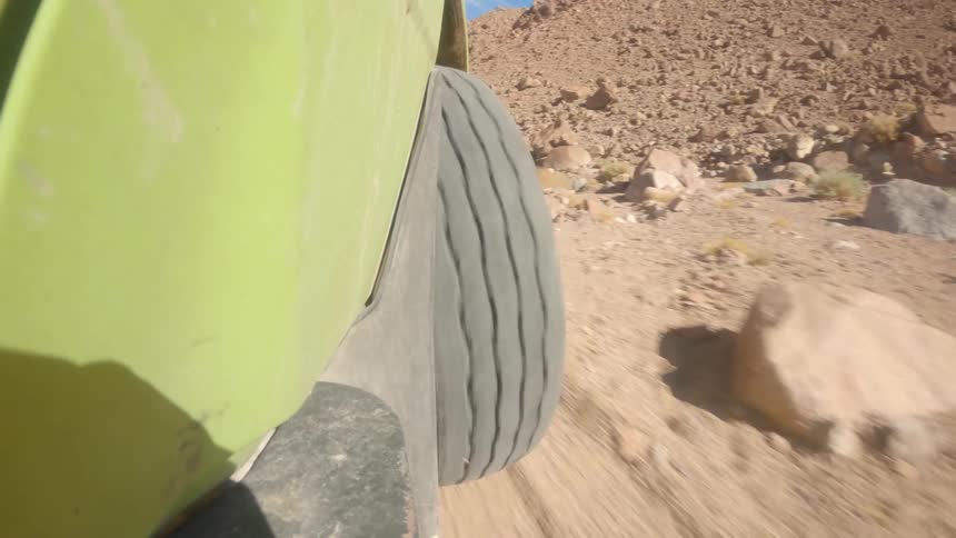 A low-angle shot from the side of an off-road vehicle shows its tire spinning as it drives along a rugged, sandy desert road in slow motion