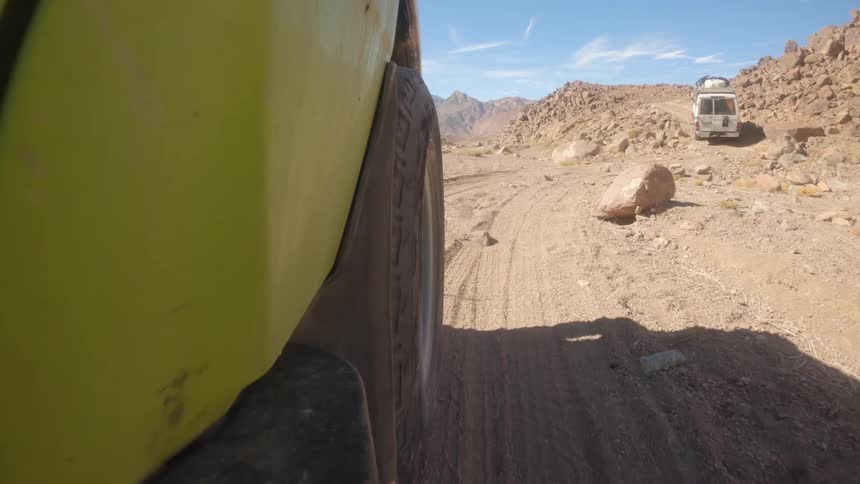 A slow-motion, low-angle shot from a green off-road vehicle shows its tire kicking up dust as it follows another camper van through a rocky desert canyon