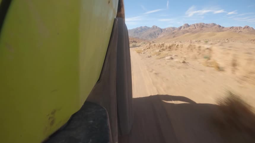 Low-angle point-of-view from a green off-road vehicle driving through a vast desert landscape with mountains on the horizon