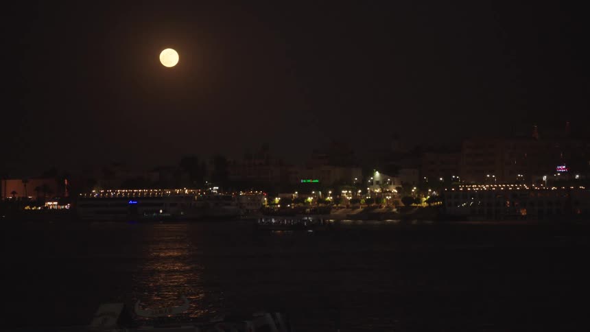 A brilliant full moon shines over the illuminated city on the banks of a river at night
