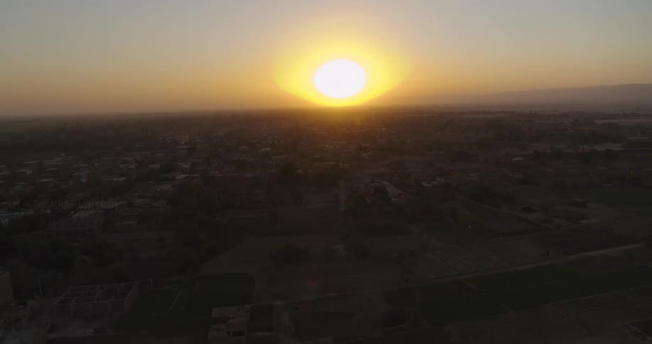 A stunning aerial pullback reveals a vast rural landscape with a village and farmlands bathed in the warm glow of a beautiful sunset