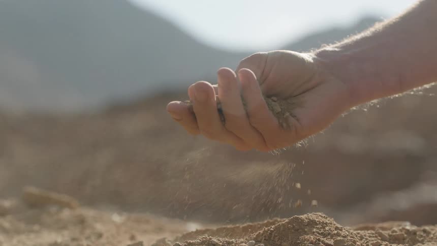 Slow motion close-up of dry earth sifting through a person's hand in a sunlit, arid environment