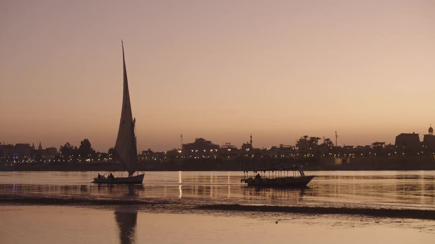 A silhouetted felucca and a tourist boat glide on the tranquil Nile River against the backdrop of an illuminated Egyptian cityscape at dusk