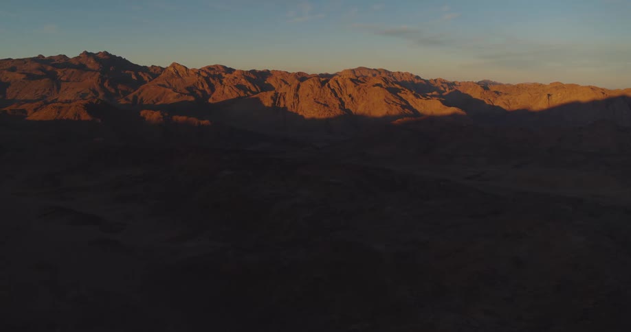 A stunning aerial view of the sun rising over a vast mountain range, casting long shadows across the desert landscape