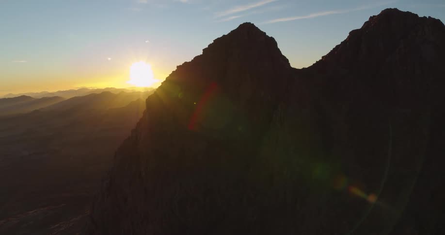 An epic aerial shot captures the sun descending behind the rugged peaks of a silhouetted mountain range at dusk
