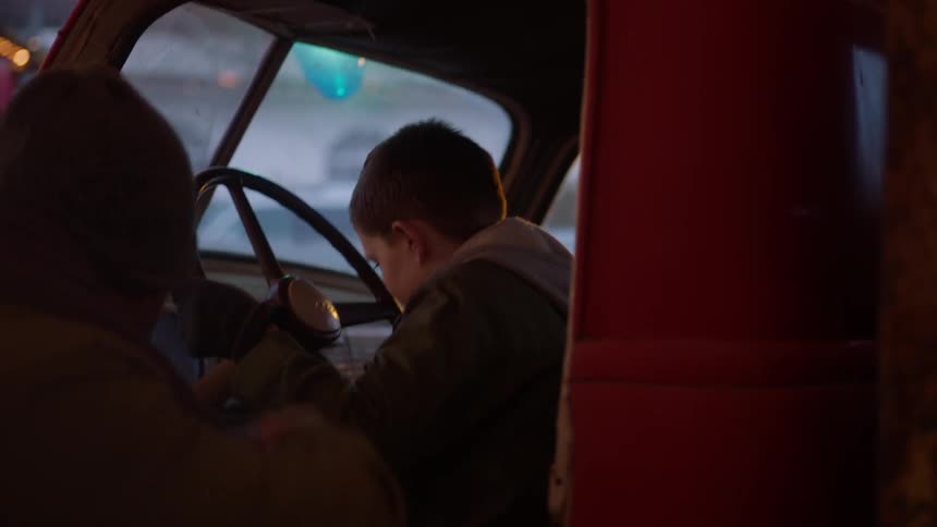 A man opens a door and lets a boy sit behind the wheel of his truck