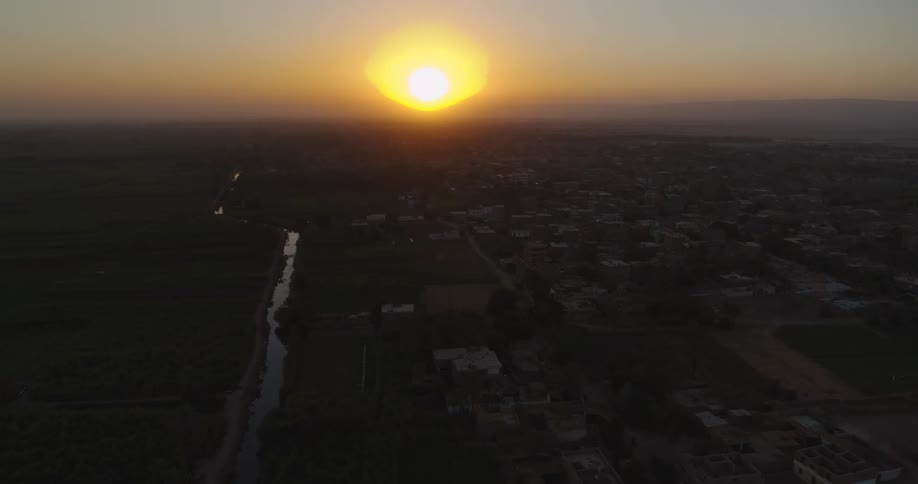 An aerial view captures the golden sun setting over a rural village, casting a warm glow on the surrounding fields and river