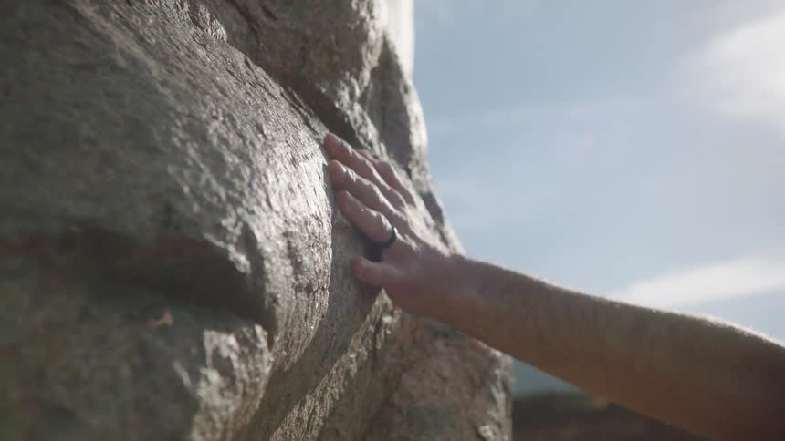 A close-up of a man's hand with a ring slowly caressing the sunlit face of a large boulder