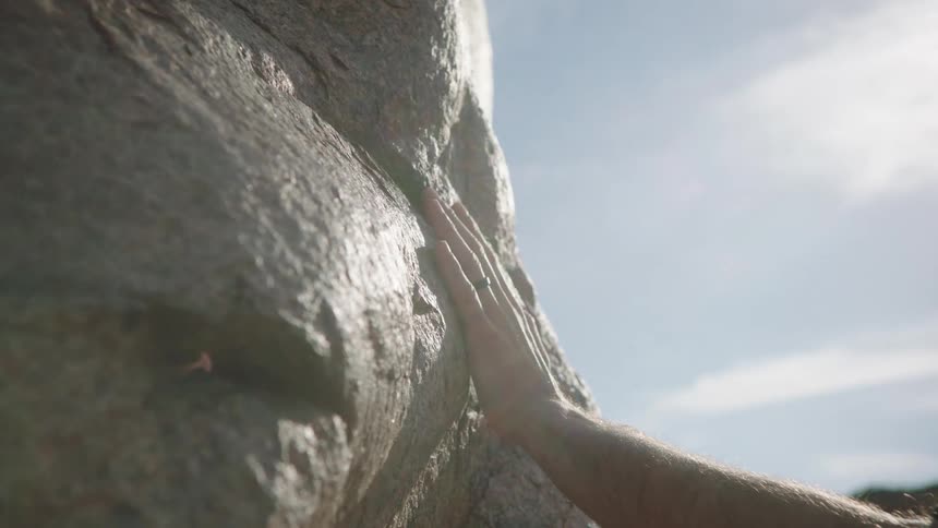 Connecting with nature, a man's hand gently touches a large sunlit boulder in slow motion