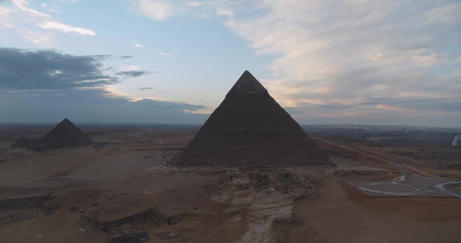 An ascending aerial shot reveals the iconic Great Pyramid of Giza standing majestically in the desert against a cloudy sunset sky