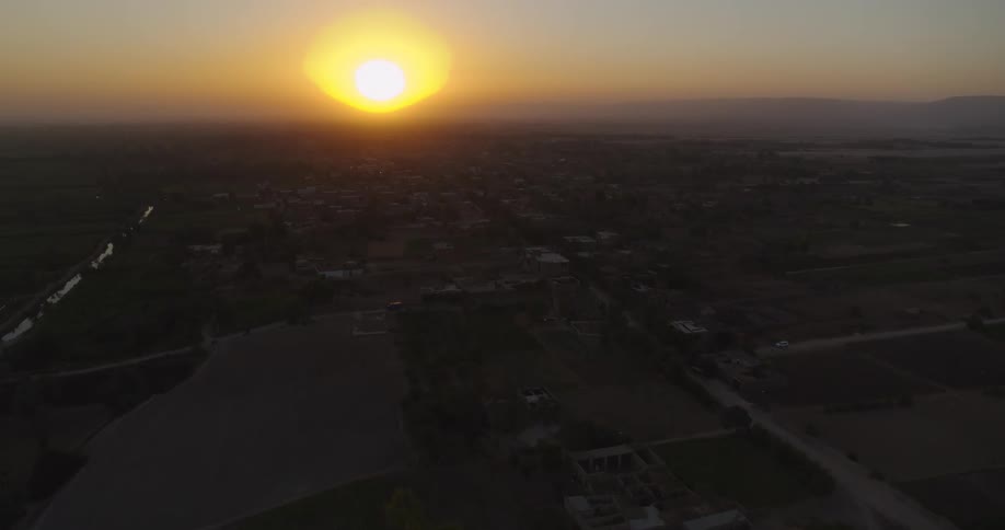 The golden sun rises over a sprawling rural village with farmlands and roads in this slow, hazy aerial shot