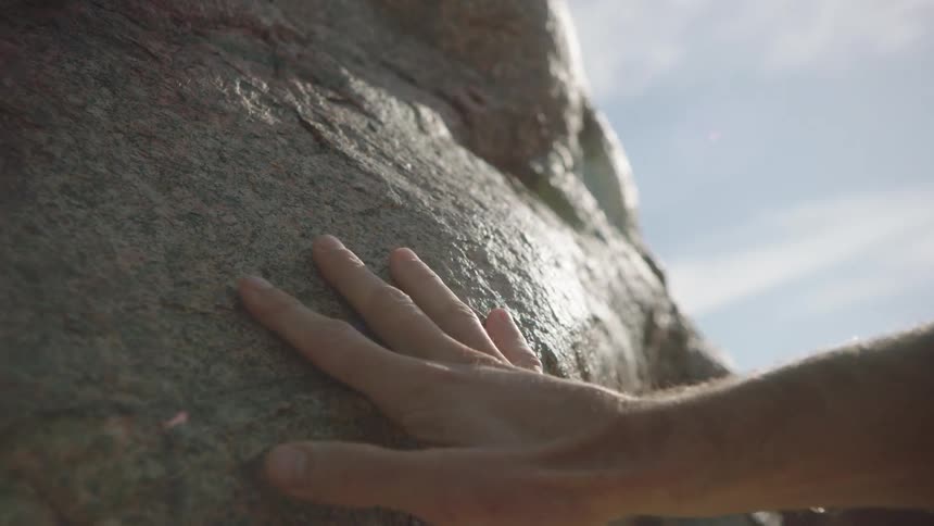 A man's hand gently glides over the smooth surface of a sun-warmed boulder in slow motion