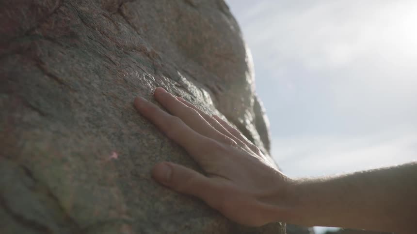 A close-up slow-motion shot shows a person's hand gently caressing a sun-drenched rock