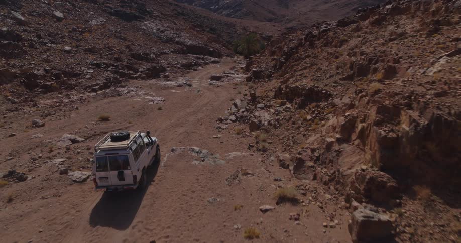 Aerial view follows a white SUV on an off-road adventure, driving down a dusty track through a remote and rocky canyon