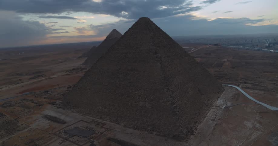 A majestic aerial pullback reveals the ancient Pyramids of Giza standing in the vast Egyptian desert under a dramatic twilight sky