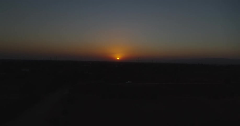 An aerial view captures the sun rising over a silhouetted rural landscape with power lines against a beautiful orange and blue sky
