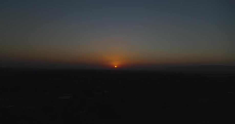 An expansive aerial view shows the sun setting on the distant horizon, casting a warm glow over a silhouetted landscape at twilight