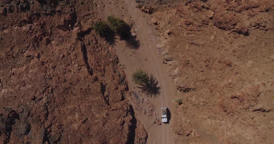 An aerial top-down view shows a white pickup truck driving on a dusty road through a rugged red rock canyon