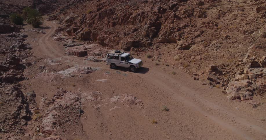 An aerial view shows a white 4x4 vehicle driving along a winding dirt road through a rugged desert canyon