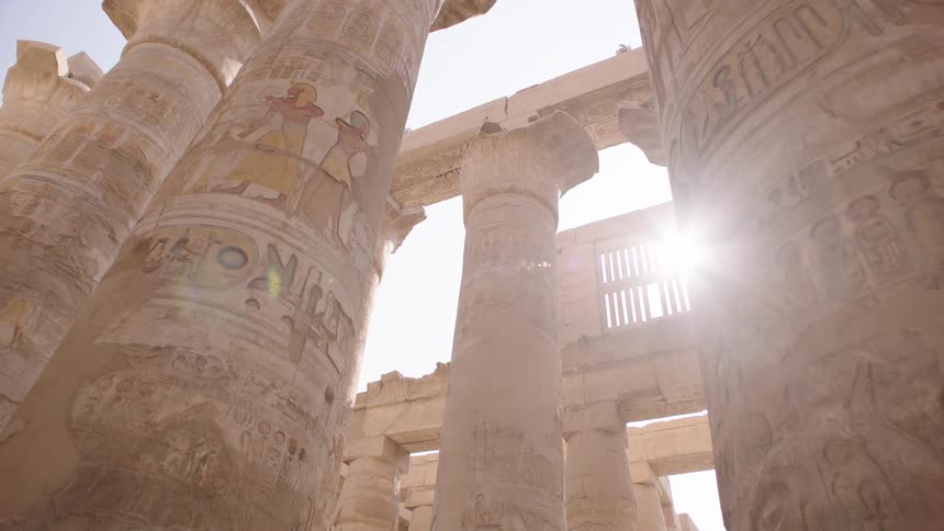 Majestic pillars with ancient Egyptian hieroglyphs and paintings stand tall as sunlight flares through the temple ruins