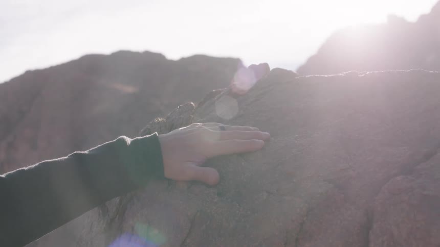 A person's hand gently glides over the surface of a sunlit boulder in a rocky landscape in slow motion