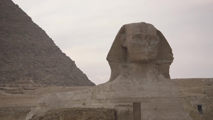A reveal shot focuses on the ancient Great Sphinx of Giza, with a majestic pyramid visible in the background against a pale sky