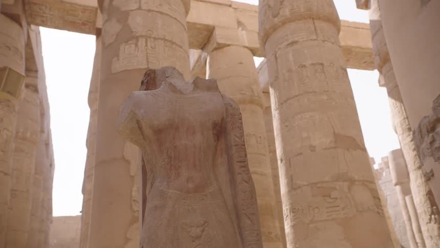A low-angle shot reveals a headless ancient Egyptian statue standing tall amongst the grand, hieroglyphic-covered columns of a temple in Luxor