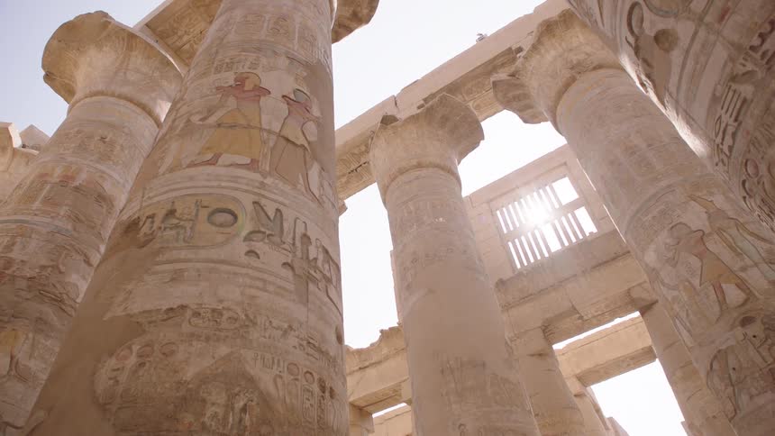 A low-angle view shows sunlight flaring through the massive, ancient pillars covered in colorful hieroglyphics at the Karnak Temple in Luxor, Egypt