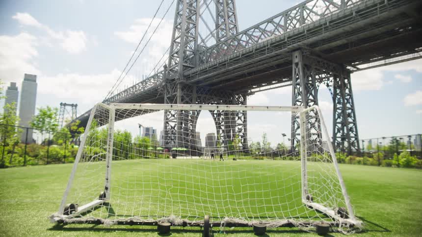A slow tilt down from the massive Williamsburg Bridge reveals a soccer goal on a sunny, urban turf field in New York City