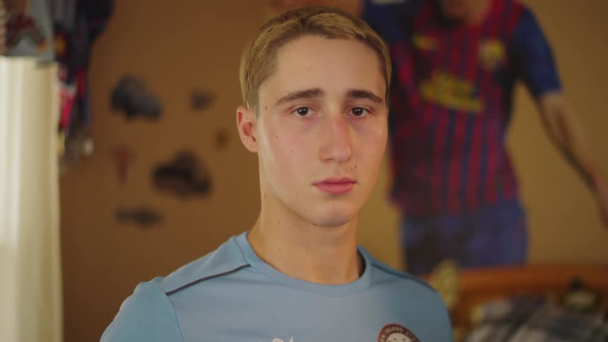 A close-up portrait of a serious young man in a soccer jersey looking at the camera in his bedroom