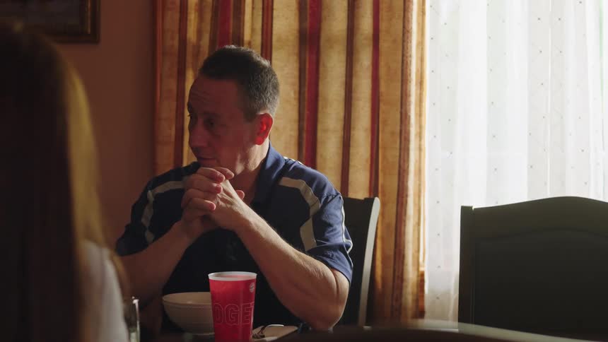 A pensive middle-aged man listens intently during a meal at the dining table, eventually breaking into a warm smile