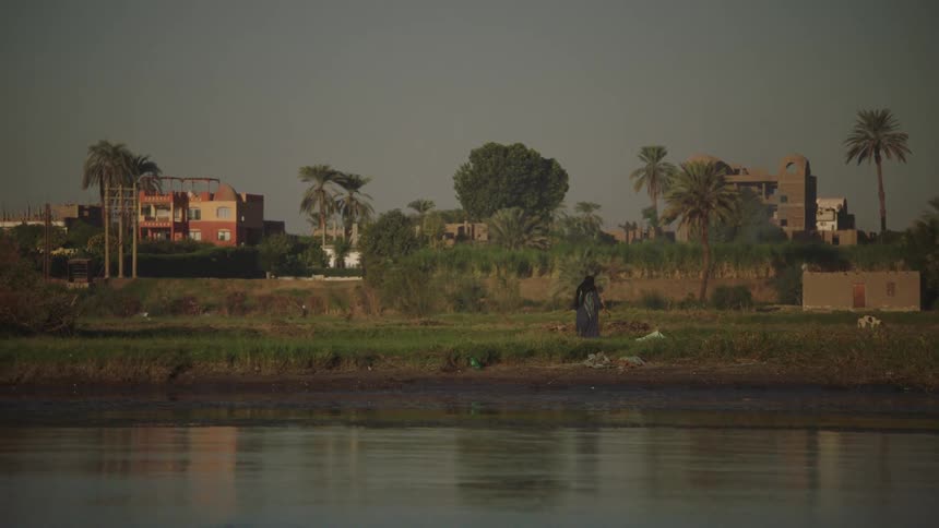 A woman in traditional robes tends to the lush green banks of the Nile River with an Egyptian village in the background