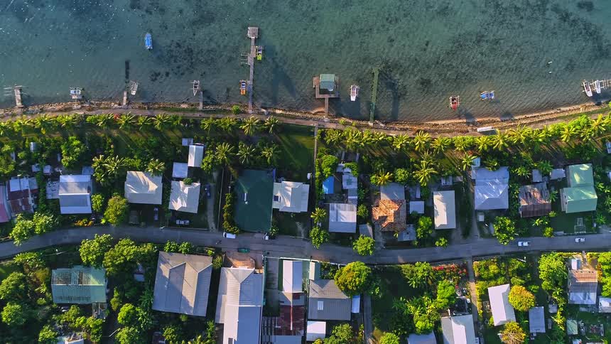 A slow top-down aerial pan over a lush tropical village situated along the clear turquoise coastline, with boats and piers dotting the shore