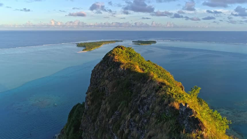 An aerial drone shot flies over a sun-kissed mountain peak, revealing a beautiful tropical island and a vast blue ocean beyond