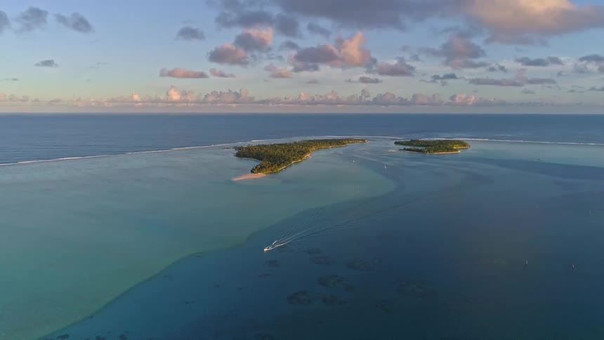 A breathtaking aerial shot captures a lone boat sailing through a tranquil turquoise lagoon towards two tropical islands at sunrise